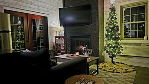 Un salon chaleureux avec une cheminée, un téléviseur fixé au-dessus, un sapin de Noël et une petite table sur laquelle est posé un jeu de société.