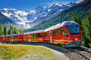 Um trem vermelho sobre trilhos, com montanhas ao fundo.