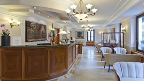 A hotel lobby with a wooden reception desk, a chandelier, and seating area with chairs. - Alimandi Vaticano (Rome)
