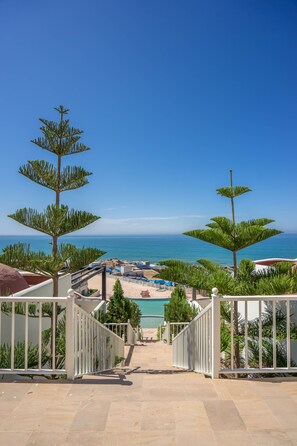 A view of a pool area with a clear blue sky and ocean in the background, surrounded by palm trees and a white fence.