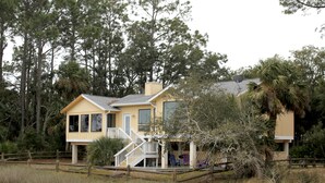 Une maison à deux étages, dotée d’un porche et d’un escalier, entourée de grands arbres et d’une clôture en bois.