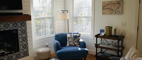 A living room with a blue armchair, a glass coffee table, a fireplace, and a bookshelf.