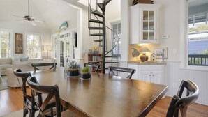 A spacious dining area with a wooden table and chairs, a spiral staircase, and a view of the outdoors through a window. - Updated in Seaside, Amazing Porches, Close to West Side Pool + 2 Adult Bikes! (Santa Rosa Beach)