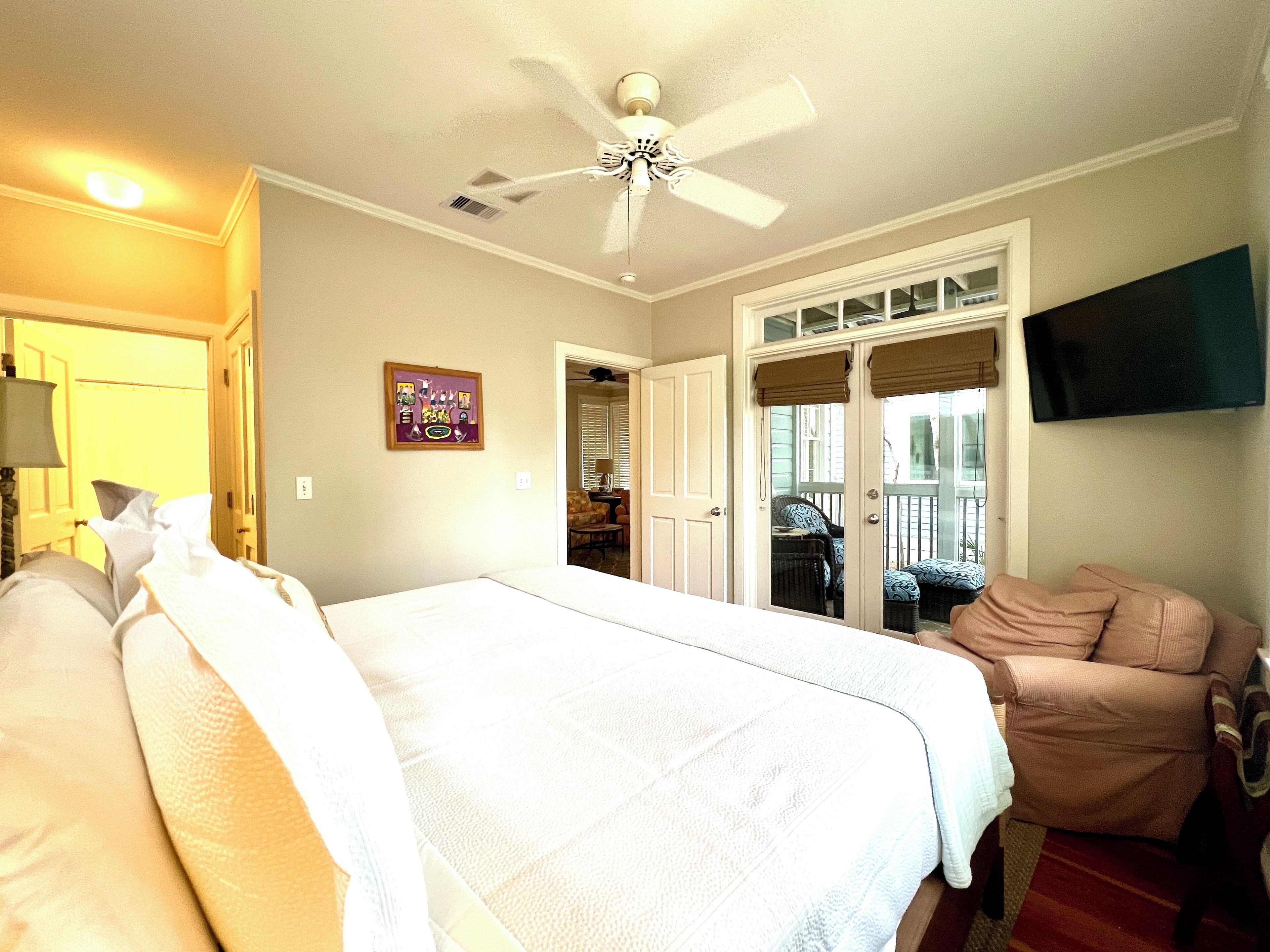 A hotel room with a large bed, a ceiling fan, a TV mounted on the wall, and a view of the outside balcony.