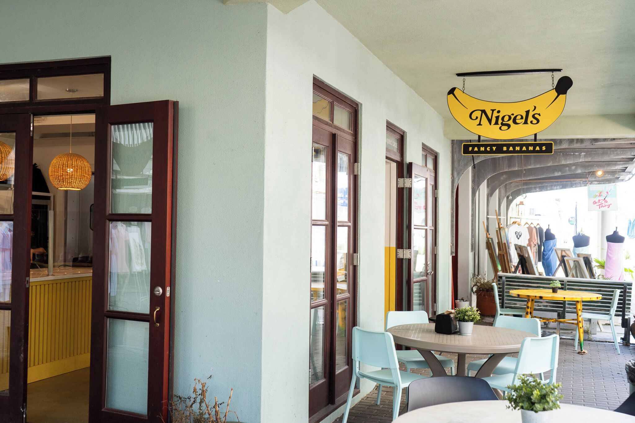 Ein Café mit einem Schild, auf dem 'Nigel's Fancy Bananas' steht, mit einem runden Tisch und Stühlen und Blick auf die Straße durch große Fenster.