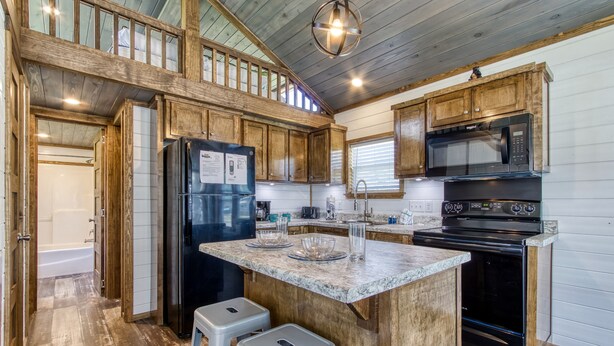 A modern kitchen with wooden cabinets, a black refrigerator, a microwave, and a stove. There are two stools and a window with blinds.