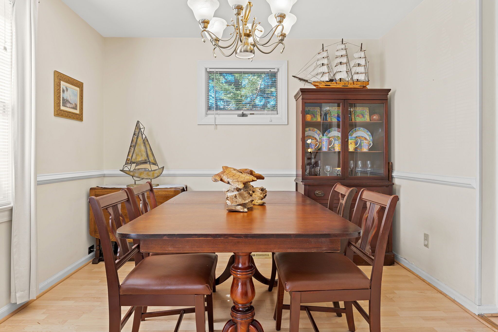 Une salle à manger avec une table en bois, des chaises, un lustre, une maquette de bateau et un buffet aux portes vitrées.