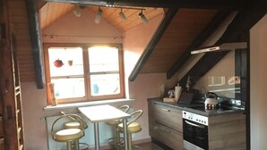 Een keuken met een centraal eiland, twee barkrukken, een raam met gordijnen, en een houten plafond.