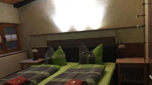 Een kamer met twee bedden, elk met beddengoed en kussens in groen en grijs. Er is een raam met luxaflex, een nachtkastje met een lamp en een houten kast.