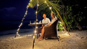 A couple seated at a table under a palm tree, with string lights and a beach view.
