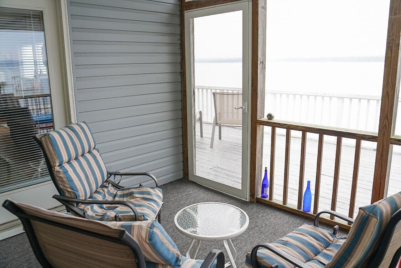 Un balcon donnant sur l’eau, une chaise à rayures, une petite table et deux bouteilles bleues.