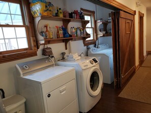 A laundry area with a front-loading washer and dryer, shelves with cleaning supplies, and a window with blinds. - Historic Hardware Inn with River Views: Suite 102 (Stockport)
