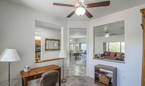 A room with a ceiling fan, a desk, a chair, a lamp, a mirror, and a view of a living area with a sofa and a window. - Sun City Grand Ranch style house (Surprise)