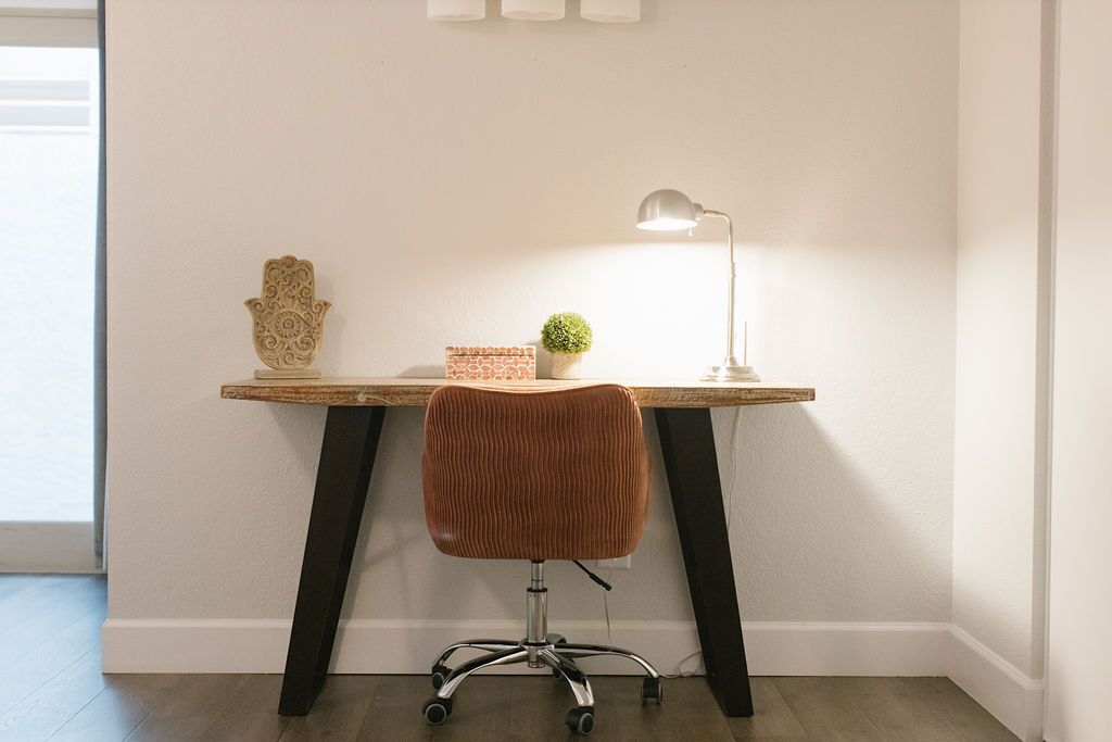 Um espaço de trabalho com escrivaninha de madeira, luminária, objeto decorativo e uma cadeira.