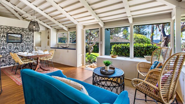 Amplia habitación con techo alto, zona de comedor con mesa y sillas, sofá azul, sillones de mimbre y vistas al jardín a través de amplias ventanas.