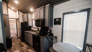 A compact kitchen with grey cabinets, a black refrigerator, a microwave, and a coffee maker. There's a round table with two chairs and a window with blinds.