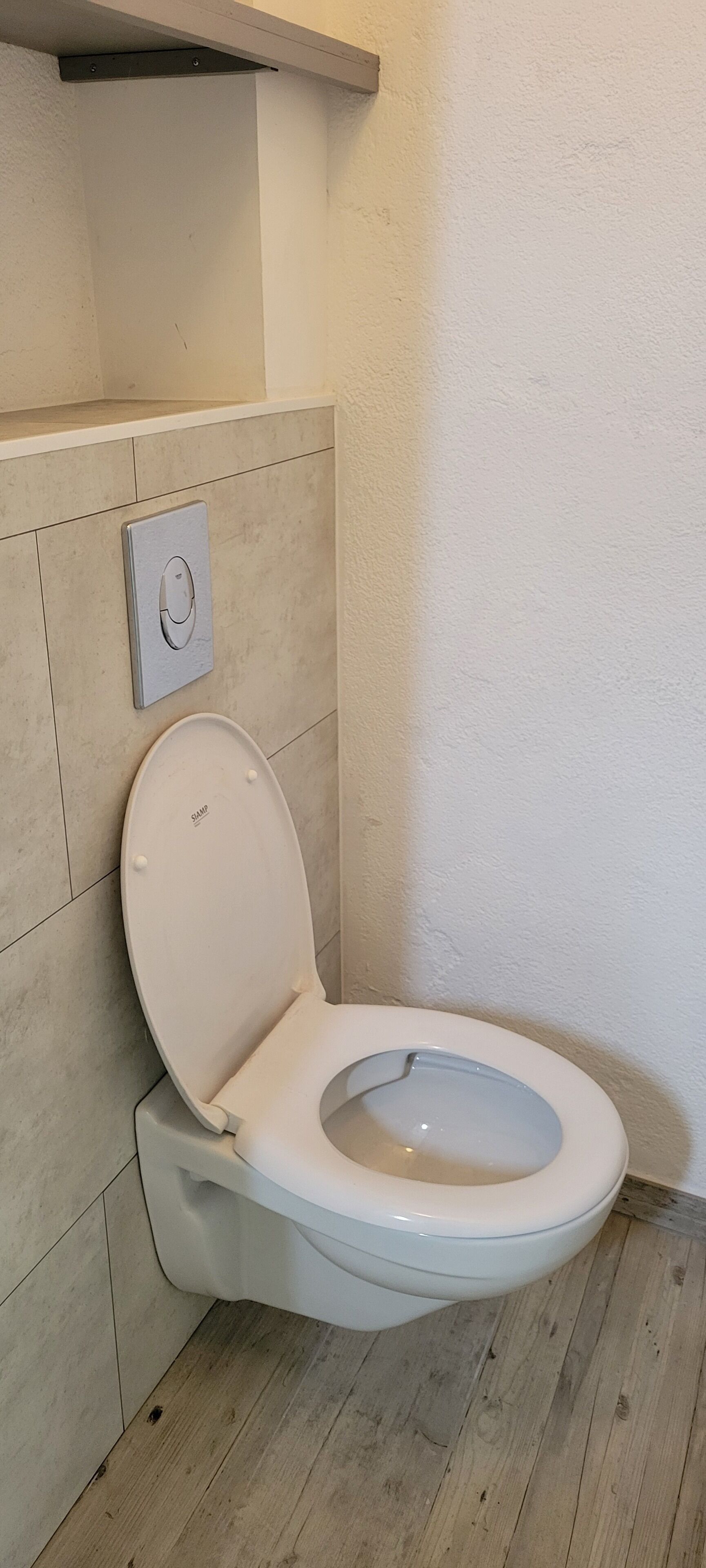 A modern bathroom with a white toilet, a wall-mounted flush handle, and a wooden floor.