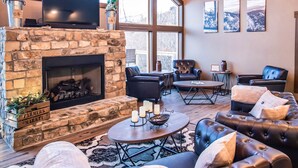 A spacious living room with a stone fireplace, leather sofas, a television, and large windows offering a view of the outdoors. - Gorgeous Luxury Cabin! 360 Views, Fire Pit, Theater Room - Prime Location!  (Gatlinburg)