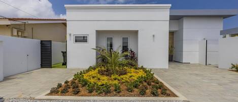 Une maison moderne à l’extérieur blanc, avec un jardin devant agrémenté de fleurs jaunes et une allée pavée.