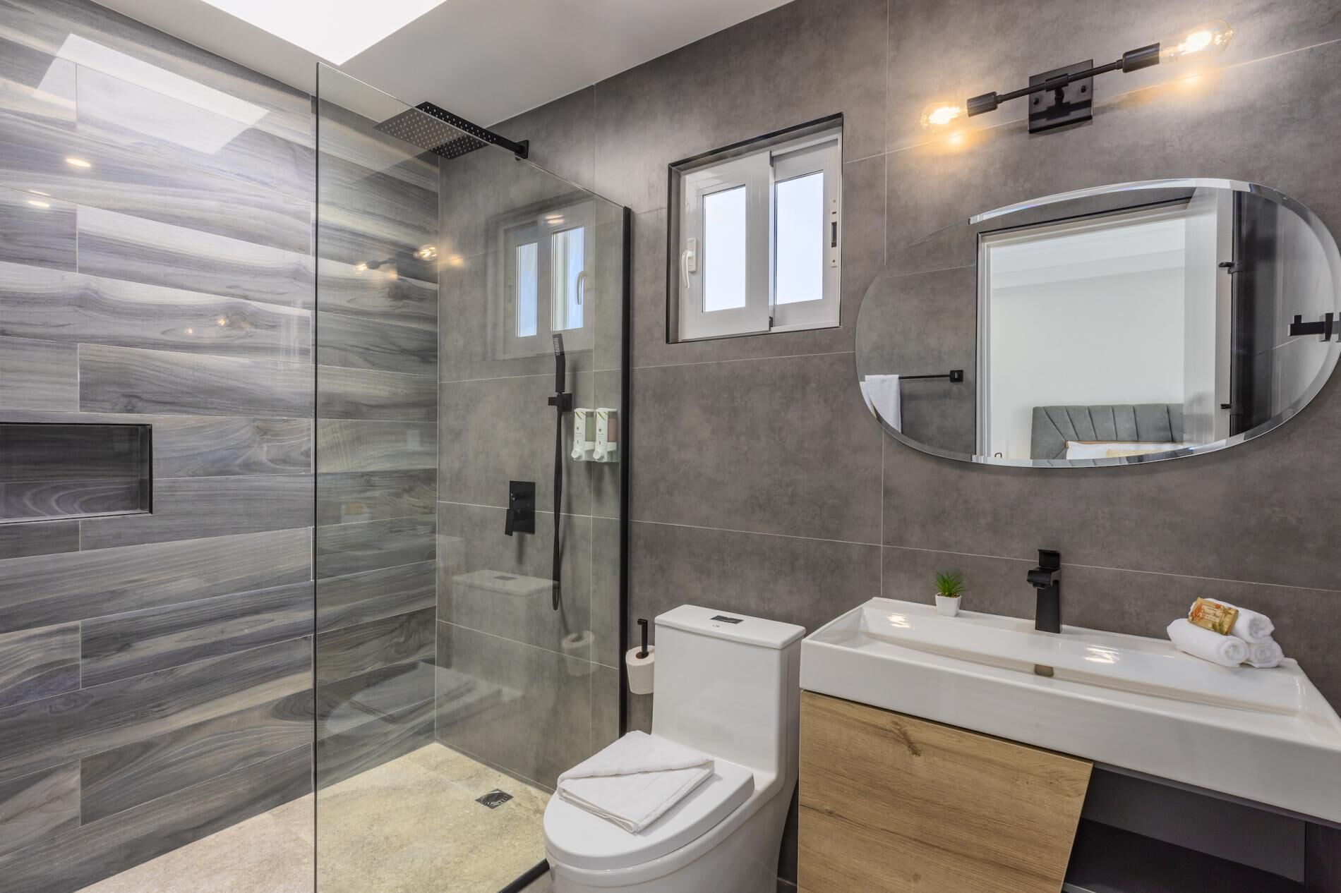 A modern bathroom with a glass-enclosed shower, a toilet, a vanity with a mirror, and a small window.