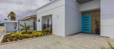 Ein modernes Gebäude mit flachem Dach, weißen Außenwänden und einer blauen Tür. Ein gepflasterter Weg und ein kleiner Garten mit verschiedenen Pflanzen sind ebenfalls vorhanden.