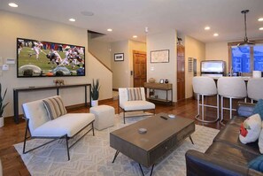 A living room with a flat-screen TV, a sofa, a coffee table, and a bar area with stools.