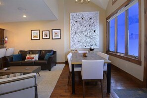 A modern living room with a sofa, coffee table, dining table, and chairs. There are framed pictures on the wall and a large abstract painting.