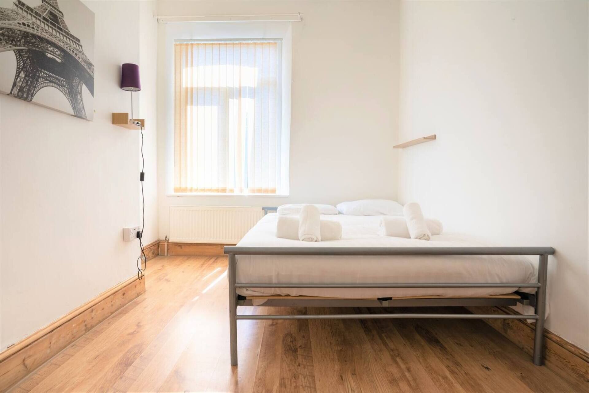 A bedroom with a bed, a window with blinds, a radiator, and a wall-mounted lamp.