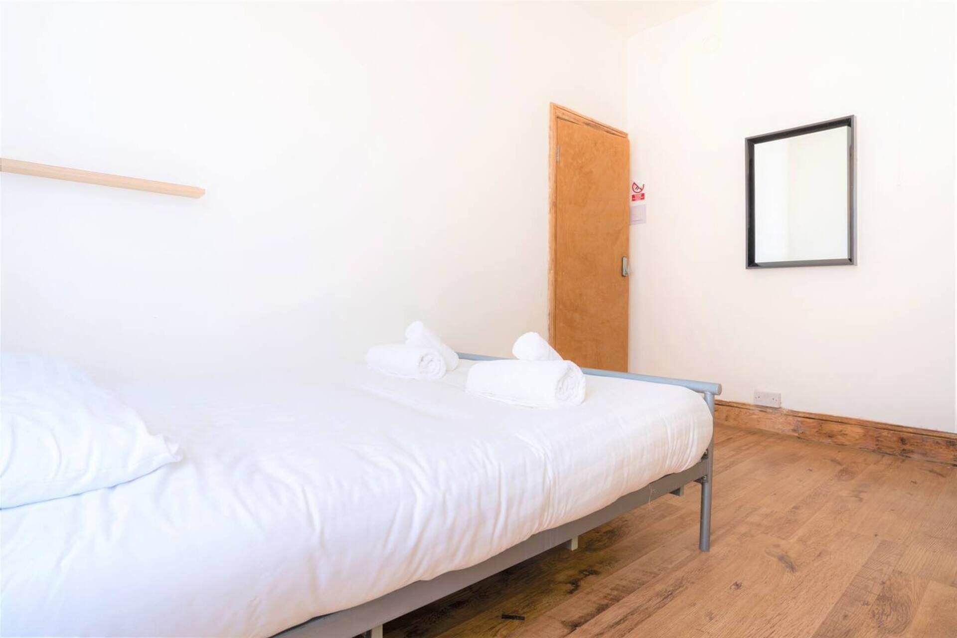 A single-bed room with a wooden floor, a wooden door, and a wall-mounted mirror.