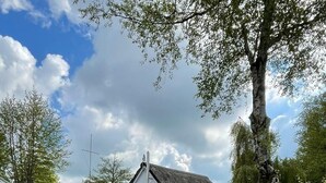 Une maison traditionnelle avec un toit de chaume, un petit jardin et une jetée en bois qui s’avance sur une étendue d’eau calme.