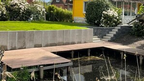 Une maison avec un toit de chaume, une petite terrasse et un étang au premier plan.