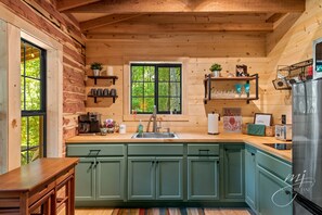 Una cocina con paredes de madera, un lavamanos y armarios. Hay repisas con objetos y una ventana con vista a árboles.