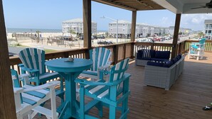 Een houten terras met turquoise Adirondack stoelen en een ronde tafel, met uitzicht op een strand en gebouwen.