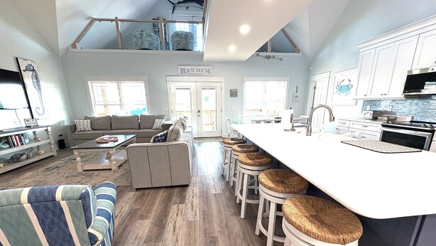 Een ruime woonkamer met een bank, een hoog plafond met dakraam, een keuken met groot kookeiland, en een bordje met de tekst 'Beach House'.