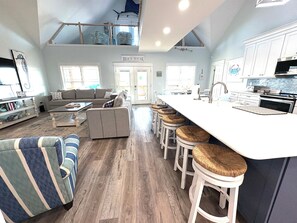 A spacious living area with a sofa, a high ceiling with a skylight, a kitchen with a large island, and a sign reading 'Beach House.'