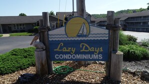 A sign for 'Lazy Days Condominiums' with a sailboat graphic, surrounded by wooden posts and greenery. - Unit B7 spacious 4bed/3bath condo perfect lakefront spot for family and friends (Osage Beach)