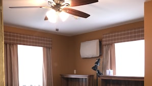 A room with two single beds, a ceiling fan, a window with curtains, a bedside table with a lamp, and a wall-mounted air conditioner. - Catskills Adventures (Roscoe)