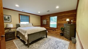 Une chambre avec un mur en bois brut, un lit avec une literie blanche, une table de chevet avec une lampe, une commode et une fenêtre avec des stores.