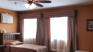 Habitación de hotel con dos camas, ventilador de techo, ventana con cortinas, una mesita y un cubo de basura.