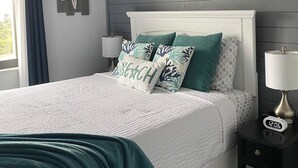 Una cama bien hecha con cabecera blanca, almohadas estampadas en blanco y verde azulado, y una manta verde azulado. Hay una mesita de noche con una lámpara y un reloj digital que marca las 12:06. Una decoración con forma de estrella de mar cuelga en la pared.