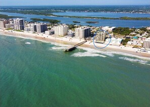 Luftaufnahme einer Küstenregion mit Hochhäusern, einer Pier, die ins Wasser führt, und freiem Blick auf Strand und Meer.