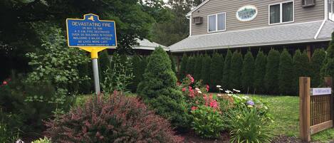 A signboard with historical information about a devastating fire, a house with a sign for Trout Town Inn, and a garden with various plants and flowers.