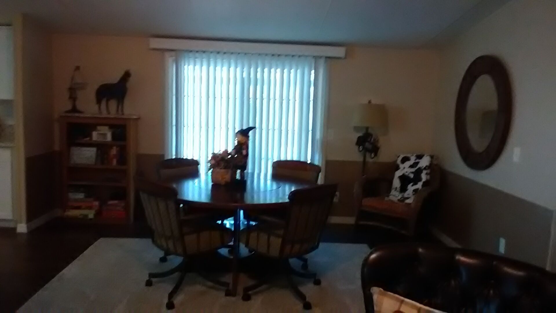 Uma sala de jantar com uma mesa redonda, cadeiras, uma estante, uma janela com persianas, um abajur e um espelho na parede.