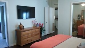 Um quarto com uma cama, cômoda, televisão na parede e um corredor que dá para outro cômodo.