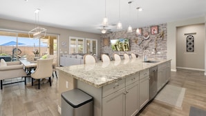 A modern kitchen with a large island, granite countertops, and stainless steel appliances. There is a dining area with a table and chairs, and a view of the outdoors through large windows. - No Fee Heated pool & Spa  gorgeous House.  (Goodyear)