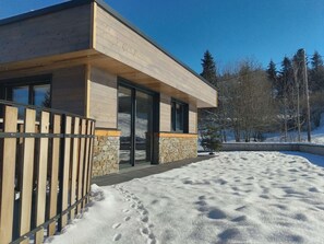 Een modern huis met stenen fundering en houten bekleding, gelegen in een besneeuwd landschap met bomen op de achtergrond.