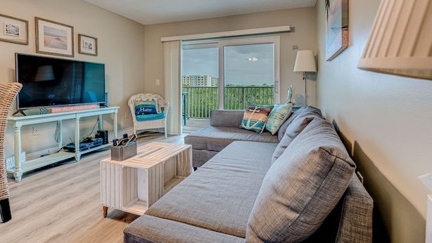 A living room with a sofa, a television, a wicker chair, and a balcony with a view of buildings.