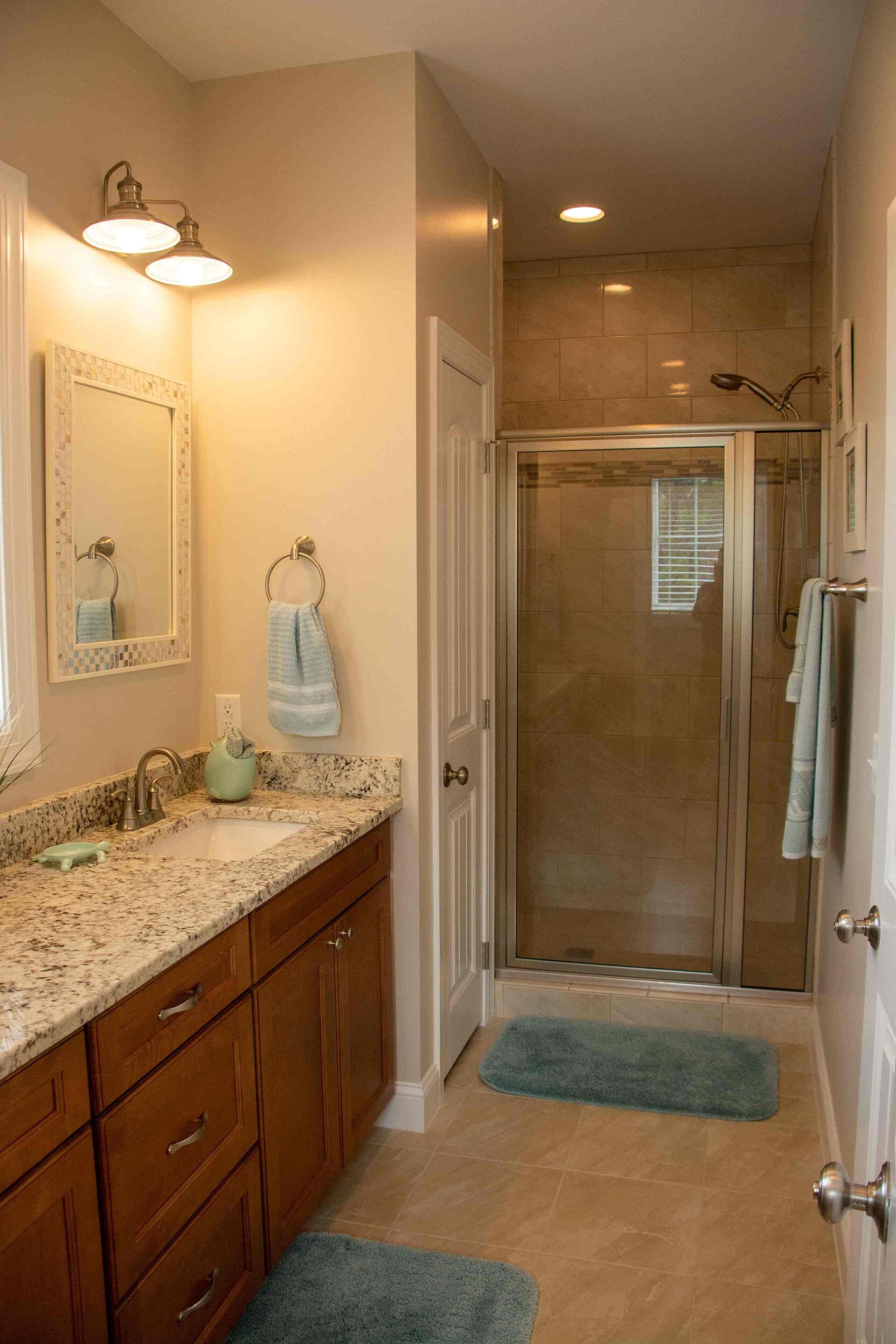 Un baño con encimera de granito, un lavabo, un espejo, un perchero para toallas, una ducha con puerta de cristal y una puerta con pomo plateado.