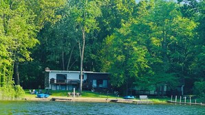 A lakeside cabin with a deck, surrounded by dense trees. - The Perfect Lake Getaway! (Paw Paw)