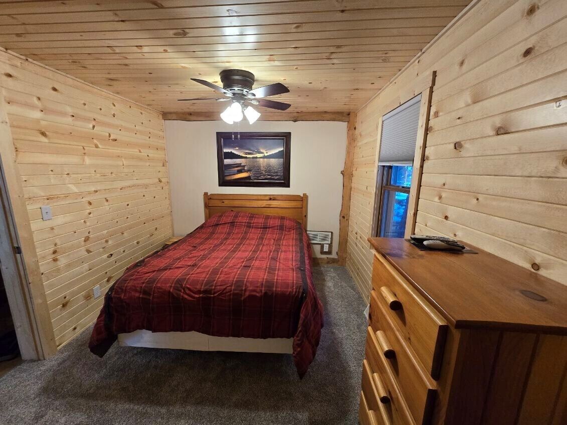 A wooden-paneled bedroom with a bed, a dresser, a ceiling fan, and a window.